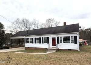 A single-story white house with black shutters and a red door, featuring a carport on the left side. The yard has patchy grass, and there are leafless trees and another house visible in the background.