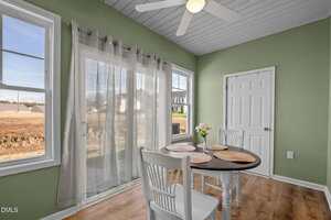 A small dining area with a round table and two white chairs is set beside large windows with sheer curtains. The walls are green, the ceiling is white with a fan, and there is a closed white door.