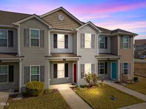 A row of two-story townhomes at 403 Winston Way in Creedmoor features beige and tan siding, multiple windows with shutters, small covered entrances, and neatly trimmed shrubs beneath a pink and purple sunset sky.