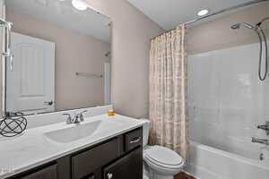 Bathroom with a white bathtub and shower, patterned shower curtain, toilet, white countertop with a sink, dark vanity cabinet, large wall mirror, and a small decorative item on the counter.