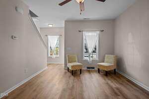 A room with light-colored walls and wood flooring features two patterned accent chairs near two windows with sheer white curtains. A ceiling fan is overhead, and stairs are visible to the left.