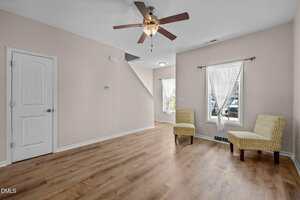A living room with light wood flooring, beige walls, a ceiling fan with lights, two yellow patterned chairs by two windows with sheer curtains, and a closed white door. Stairs are visible in the background.