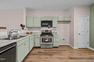 A kitchen with light green cabinets, stainless steel appliances, marble countertops, a double sink, light wood flooring, and light pink and green walls. A white door is on the right.