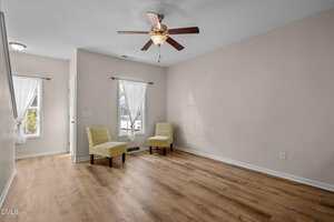 A mostly empty living room with light wood flooring, beige walls, two windows with sheer white curtains, two patterned yellow chairs, and a ceiling fan with a light.