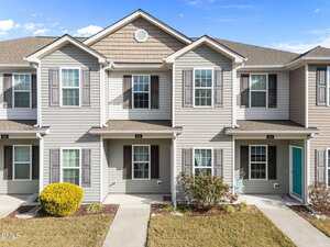 A two-story townhouse with light beige siding, brown shutters, and a small covered porch. There are trimmed shrubs and a grass lawn in front, with a clear blue sky overhead.