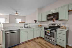 A modern kitchen with mint green cabinets, stainless steel appliances, a double sink, light countertops, and wood flooring. The space is bright with natural light from the adjacent dining area.