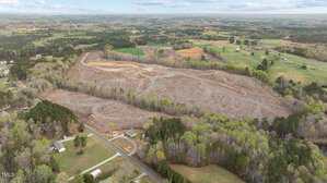 Aerial view of a large, cleared parcel of land surrounded by trees and rural properties, with winding dirt paths visible across the cleared area. Scattered houses and farmland are seen in the background.