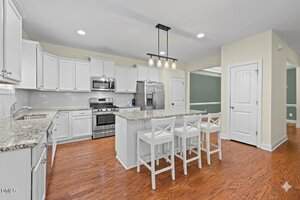 Modern kitchen with white cabinets, stainless steel appliances, granite countertops, a central island with three white barstools, pendant lighting, and hardwood floors. A green accent wall is visible in the background.