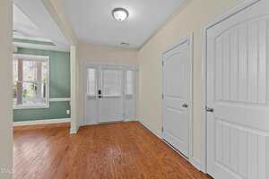 A foyer with wood flooring, beige and green walls, white trim, and two closed white doors. The entry has double front doors with glass panels and side windows, and a chandelier-style ceiling light.