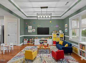 A playroom with green walls, a colorful rug, and various toys. There are yellow and red kid-sized chairs, a blue sofa, a TV on the wall, shelves with bins, a small white table, and wall decals of stars and planets.