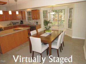 A digitally staged kitchen and dining area with wood cabinets, an island, stainless steel appliances, a wooden dining table with six white chairs, and large windows. The text "Virtually Staged" appears at the bottom.