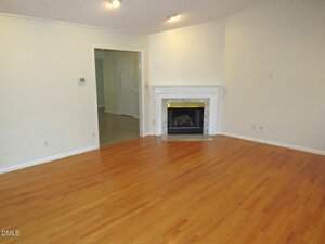 Empty living room with hardwood floors, white walls, a white fireplace with a marble surround, and a wide doorway leading to another room with tile flooring. Ceiling spotlights are turned on.