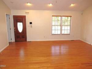 An empty room with light-colored walls, a wooden front door with a glass oval, double windows with beige blinds, and hardwood floors. Ceiling lights are on, and there are vents on the floor.