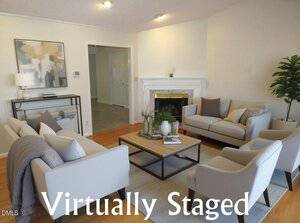 A living room with a light color scheme, featuring a fireplace, two sofas, two armchairs, a coffee table with decor, a potted plant, a console table, wall art, and the text "Virtually Staged" at the bottom.