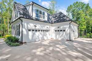A white two-story house with black roof shingles features three garage doors and a concrete driveway, surrounded by trees and greenery under a blue sky.