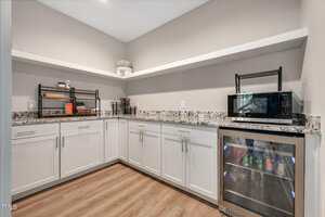 A kitchenette with white cabinets, granite countertops, light wood flooring, open shelves with decorative items, a microwave, and a glass-front mini fridge stocked with drinks.