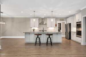 A modern kitchen with white cabinets, stainless steel appliances, a large island with granite countertop, two barstools, pendant lights, and wood flooring. The space is open and well-lit.