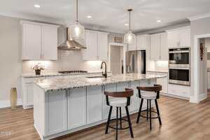 A modern kitchen with white cabinets, granite countertops, a center island with two chairs, stainless steel appliances including a double oven, and pendant lights hanging above the island.