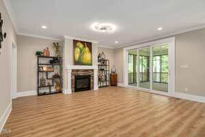 A spacious living room with light wood flooring, a white fireplace with stone accents, shelves on each side, a large painting above the mantel, and sliding glass doors leading to a screened porch with trees visible outside.