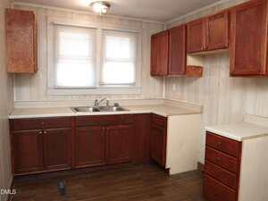A small kitchen with dark wood lower cabinets, matching upper cabinets, light countertops, a stainless steel sink under two windows with blinds, and wood-patterned flooring. Walls are light-colored with a subtle pattern.