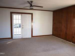 An empty room with beige carpet, white walls on two sides, wood paneling on one wall, a white door with glass panels, a ceiling fan, and a window letting in natural light.