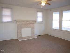 A living room with beige carpet, white walls, a white brick fireplace, two small windows with blinds above the fireplace, a ceiling fan with a light, and two large windows on the right wall.