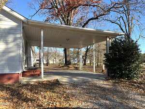 A single-car carport attached to the side of a white house with red brick foundation, surrounded by leafless trees, a bush, and scattered fallen leaves on the ground. The sky is clear and blue.
