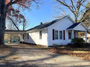 Single-story white house with dark shutters, an attached carport on the left, gravel driveway, small front porch, and leafless trees in the yard. The sky is clear and blue.