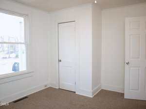 A small empty room with white walls, a beige carpet, a window on the left, a closed door in the center, and another door partially open on the right. Natural light comes through the window.