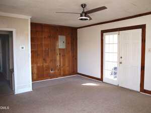 A simple room with beige carpet, wood-paneled wall, white-painted walls, ceiling fan, a door with glass panes, and a small hallway to the left. The room is empty and well-lit by natural light.