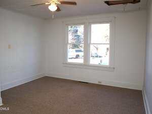 Empty room with beige carpet, white walls, a ceiling fan with light, two windows, and a hole in the ceiling near the corner. Sunlight is coming through the windows, and a vehicle is visible outside.