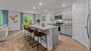 A modern kitchen with light gray cabinets, stainless steel appliances, and a central island with three brown barstools. The room features wood flooring and a living area with a beige sofa and blue curtains in the background.