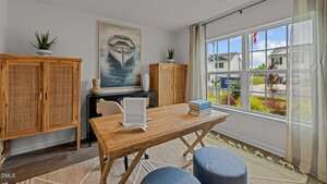 A bright home office with a wooden desk, two blue ottomans, two wooden cabinets, a painting of a boat on the wall, books on the desk, and a large window overlooking a suburban street.