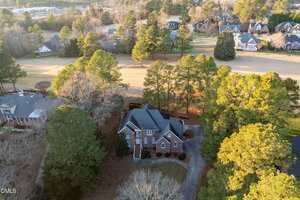 Aerial view of a two-story brick house surrounded by trees, with a curved driveway and a golf course or open grassy area behind it; neighboring houses and roads are visible in the background.
