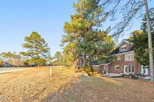 A two-story brick house with white-framed windows sits next to tall pine trees on a grassy, slightly sloped yard under a clear blue sky. A paved driveway is partially visible on the left.