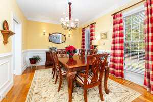 A formal dining room with a wooden table and eight chairs, a red poinsettia centerpiece, large windows with red plaid curtains, a chandelier, and a mirror above a sideboard against a yellow wall.