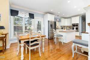 Bright, spacious kitchen with white cabinets, stainless steel appliances, a central island with stools, and a wooden dining table set for two next to large windows with blinds and valances. Hardwood floors throughout.