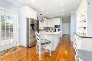 A bright, modern kitchen with white cabinets, stainless steel appliances, a center island with a gray cushioned chair, hardwood floors, and a hutch with white dishes on open shelves. Natural light enters through a glass door.