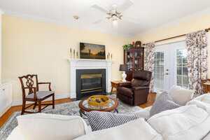 A living room with a white fireplace, TV above it, armchair, wooden chair, round coffee table, beige sofa, glass-door cabinet, lamp, and patterned curtains framing French doors. Walls are light yellow and floor is wood.