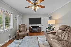 A living room with two brown leather chairs, a brown sofa, a ceiling fan with lights, a large TV on a stand, a table, a lamp, and two windows with blinds. The floor is hardwood and walls are light gray.