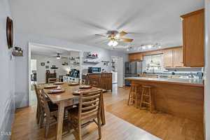 Open kitchen and dining area with wooden floors, a ceiling fan, wooden cabinets, a stainless steel refrigerator, countertop with stools, and a dining table set for four. The space is bright and organized.