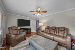 A living room with a brown leather recliner and matching sofa, a large ottoman-style coffee table, a TV on a wooden console, a floor lamp, a ceiling fan, and light gray walls with white trim.