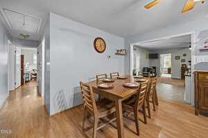 A dining area with a wooden table set for six, wooden chairs, a wall clock, and light wood flooring. The room connects to a hallway, living area, and kitchen space, all with neutral-colored walls and decor.