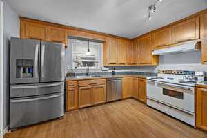 Modern kitchen with stainless steel refrigerator, dishwasher, and sink under a window. Wooden cabinets and drawers line the walls. There is a white stove and oven, and wood flooring throughout the space.