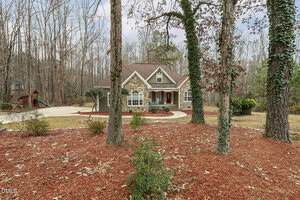 Nestled among tall trees at 1119 Corrina Road, Wake Forest, this single-story home features a gabled roof, arched window, charming front porch, mulched landscaping, and a curving driveway leading to a wooden playset in the backyard.