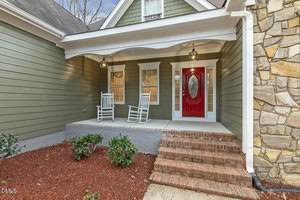 A front porch of a house with green siding, a red front door with an oval glass panel, two white rocking chairs, brick steps, mulch and shrubs along the edge, and stone accents on the right wall.