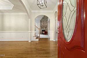 A red front door opens to a hallway with light-colored walls, wood flooring, white trim, and an archway leading to a living area with a stone fireplace and a floral arrangement.