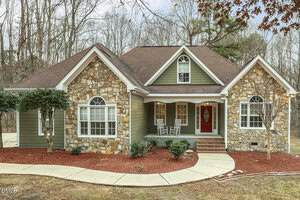 Single-story house with stone and olive green siding, a brown shingle roof, two front gables, white trim, a covered porch with two white rocking chairs, and a red front door, surrounded by mulch and trees.