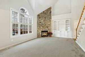 A living room with light carpet, a stone fireplace, built-in white shelves, a staircase with wooden railing, large windows with white blinds, and a vaulted ceiling. A potted plant sits beside the fireplace.