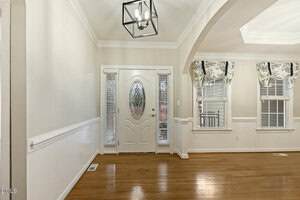 A front entryway with a white door featuring an oval glass panel and two narrow side windows, adjacent to a room with hardwood floors, cream walls, and two windows with patterned valances.